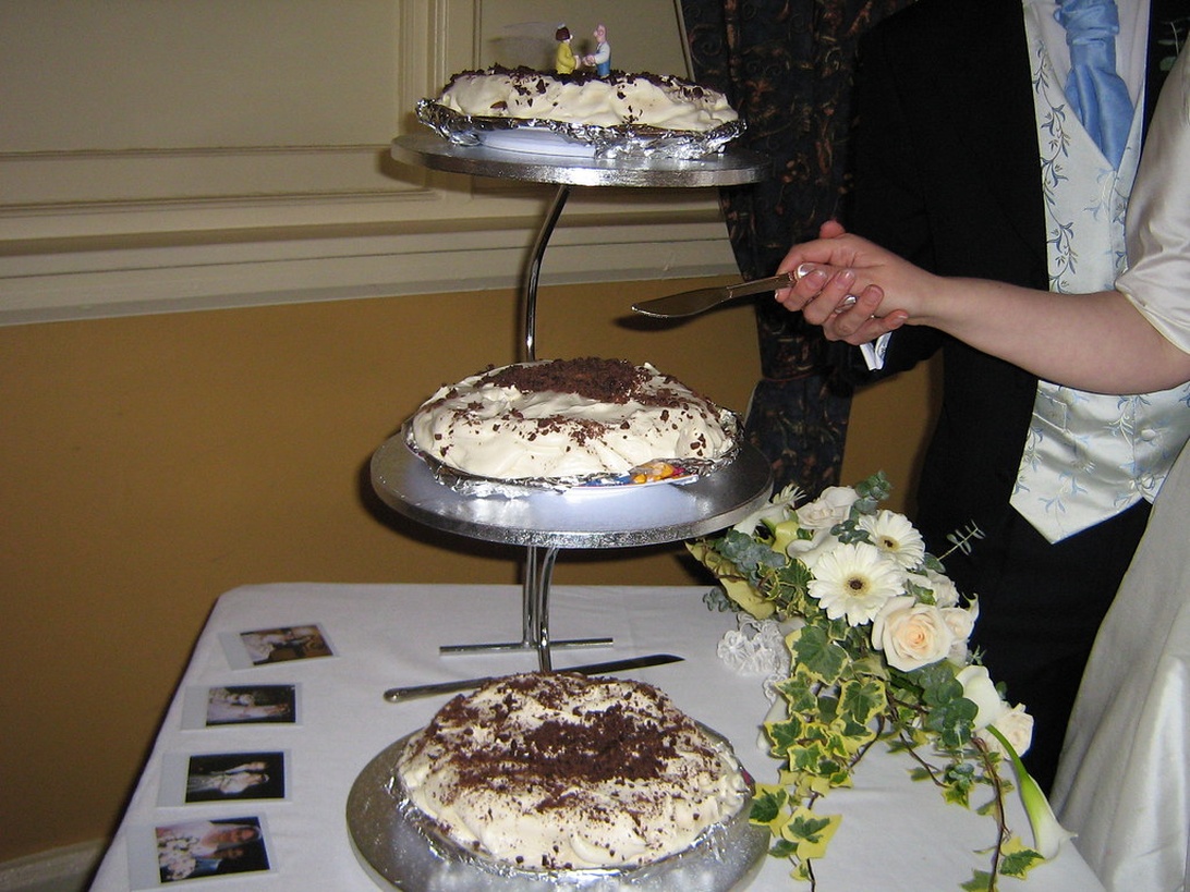 En hembakad bröllopstårta i tre våningar med vit frosting, chokladströssel och ett brudpar på toppen, där en hand syns skära upp tårtan vid ett festbord.