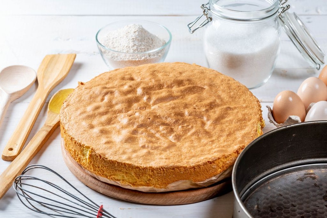 En nybakad sockerkaka på en träskärbräda, omgiven av ingredienser som ägg, mjöl och socker.