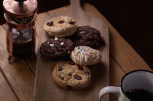 Fem olika hembakade kakor upplagda på en skärbräda i trä bredvid en kaffepress.